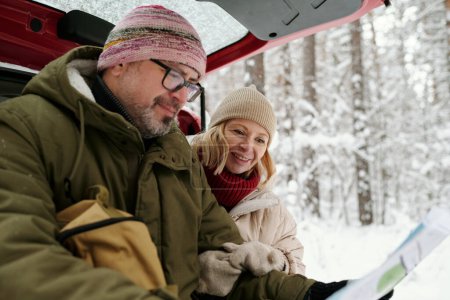 Téléchargez les photos : Couple âgé en vêtements d'hiver chauds regardant la carte papier tenue par l'homme tandis que les deux assis au coffre de la voiture contre un groupe d'arbres couverts de neige - en image libre de droit