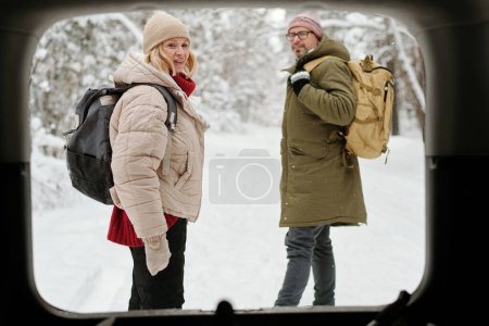 Photo for View from car trunk of mature couple of backpackers standing on road covered with snow surrounded by pine trees in the forest - Royalty Free Image