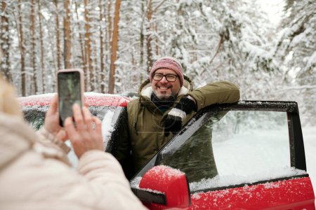 Foto de Hombre maduro feliz en gafas y ropa de invierno de pie junto a la puerta abierta del coche en el bosque mientras posando para su esposa sosteniendo el teléfono inteligente - Imagen libre de derechos