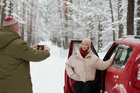Foto de Mujer madura sonriente en ropa de invierno de pie junto a la puerta abierta del coche y posando para su marido con teléfono inteligente tomar una foto de ella - Imagen libre de derechos