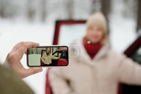 Foto de Enfoque selectivo en la mano del hombre maduro sosteniendo el teléfono inteligente mientras toma la foto de su esposa en ropa de invierno de pie junto a la puerta abierta del coche - Imagen libre de derechos