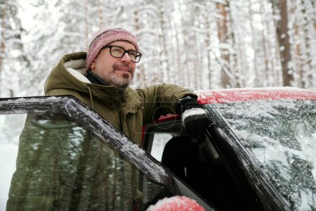Photo for Smiling mature man in eyeglasses and winterwear standing by door of his car and enjoying winter vacation in the forest covered with snow - Royalty Free Image