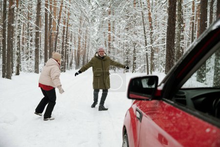 Photo for Playful mature couple in winterwear running along road in pinetree forest not far from their car while enjoying winter vacation or weekend - Royalty Free Image