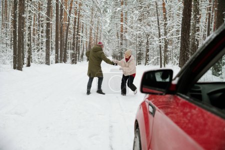 Photo for Mature man and woman playing with one another on road in pinetree forest covered with snow and enjoying their trip to the country - Royalty Free Image