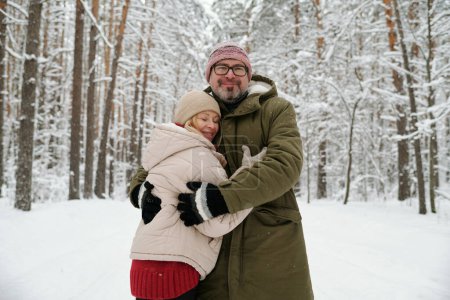 Téléchargez les photos : Heureux homme âgé embrassant sa femme dans une veste d'hiver chaude, bonnet et gants alors que les deux debout devant la caméra dans la forêt - en image libre de droit