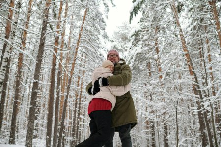 Foto de Pareja madura cariñosa en ropa de invierno de pie en el abrazo entre los pinos cubiertos de nieve mientras disfruta de fin de semana de invierno - Imagen libre de derechos