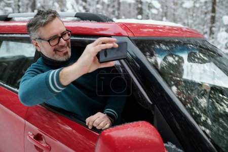 Téléchargez les photos : Homme mûr joyeux avec téléphone portable prenant selfie tout en sortant de la fenêtre de la voiture pendant les voyages d'hiver dans l'environnement naturel - en image libre de droit