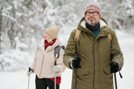 Photo for Mature man in winterwear strolling with trekking sticks in the forest covered with snow while his wife standing on background - Royalty Free Image