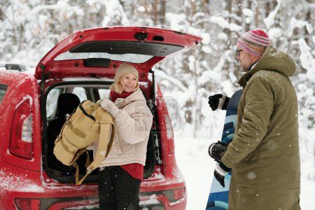 Photo for Happy senior woman with backpack looking at her husband with snowboard while taking baggage out of car trunk - Royalty Free Image