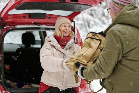 Photo for Mature cheerful woman in warm winter jacket and beanie hat passing backpack with provision to her husband while standing by car - Royalty Free Image