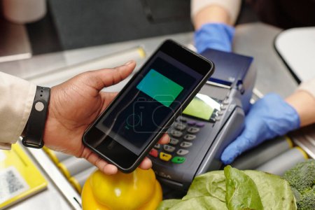 Photo for Close-up of consumer hand holding smartphone over card reader during contactless payment for food products in supermarket - Royalty Free Image