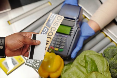 Foto de Visión general de la mano del consumidor con tarjeta de crédito y cupón de descuento sobre el terminal de pago en poder del cajero en guantes durante la transacción - Imagen libre de derechos