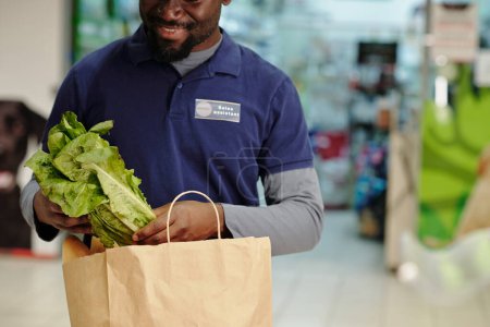 Foto de Primer plano del joven afroamericano asistente de ventas en uniforme azul poniendo verduras en bolsa de papel mientras ayuda a los clientes - Imagen libre de derechos