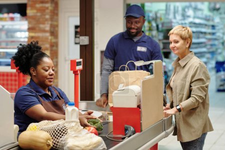 Foto de Cliente femenino rubio maduro esperando a cajero afroamericano por mostrador de compra escaneando productos alimenticios en el supermercado - Imagen libre de derechos