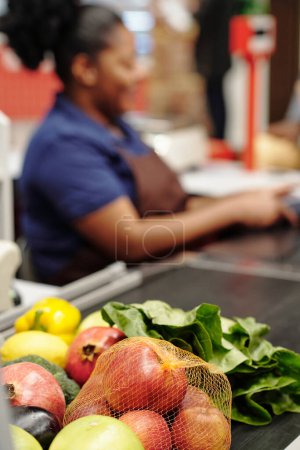 Téléchargez les photos : Focus sur les fruits et légumes frais choisis par le consommateur à la caisse ligne sur fond de jeune caissière afro-américaine - en image libre de droit