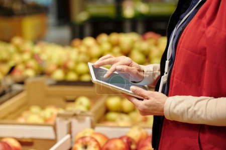 Téléchargez les photos : Gros plan d'une administratrice mature d'un supermarché en uniforme rouge utilisant une tablette tout en regardant la liste des produits pendant la révision - en image libre de droit