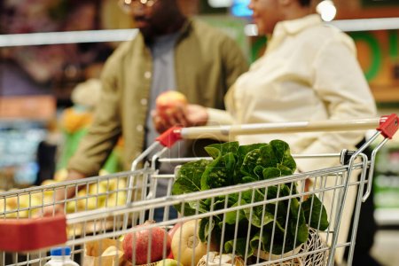 Shopping cart with fresh fruits and vegetables against young couple choosing apples while standing by display in grocery store