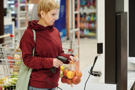 Foto de Consumidor femenino maduro rubio en ropa casual mirando a la pantalla con el precio del producto mientras escanea el paquete de manzanas maduras frescas en el supermercado - Imagen libre de derechos