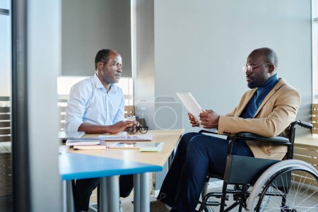 Téléchargez les photos : Jeune économiste masculin handicapé faisant rapport au patron afro-américain mature assis sur son lieu de travail devant un subordonné - en image libre de droit