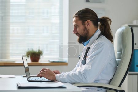Téléchargez les photos : Vue latérale d'un jeune médecin généraliste masculin sérieux consultant des patients en ligne dans un bureau clinique assis à côté d'un bureau devant un ordinateur portable - en image libre de droit