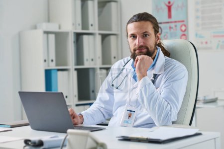 Téléchargez les photos : Jeune clinicien de sexe masculin en blouse de laboratoire assis sur le lieu de travail devant un ordinateur portable dans un bureau médical et regardant la caméra - en image libre de droit