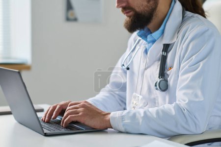 Téléchargez les photos : Gros plan d'un jeune médecin ou d'un autre spécialiste avec stéthoscope dactylographié sur un clavier d'ordinateur portable assis sur le lieu de travail - en image libre de droit