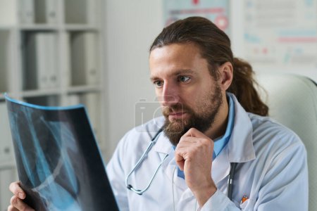 Téléchargez les photos : Radiologiste masculin sérieux tenant une image radiographique des poumons du patient devant lui alors qu'il était assis dans un cabinet médical et pensant au diagnostic - en image libre de droit