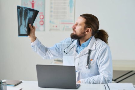 Téléchargez les photos : Clinicien masculin en blouse de laboratoire regardant attentivement la radiographie pulmonaire du patient en ligne assis près de son lieu de travail devant un ordinateur portable pendant la consultation - en image libre de droit