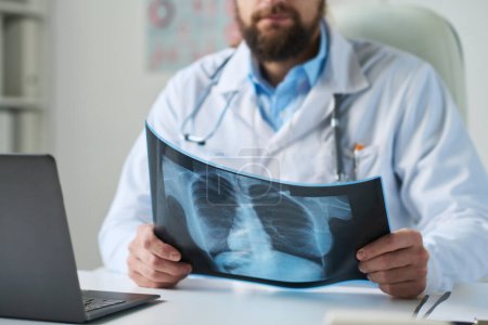 Téléchargez les photos : Gros plan du radiologue masculin barbu tenant la radiographie pulmonaire du patient en ligne tout en faisant un diagnostic par son lieu de travail devant un ordinateur portable - en image libre de droit