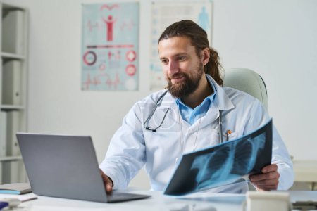Téléchargez les photos : Jeune médecin généraliste souriant avec une image radiographique regardant un patient en ligne sur un écran d'ordinateur portable pendant une consultation médicale - en image libre de droit