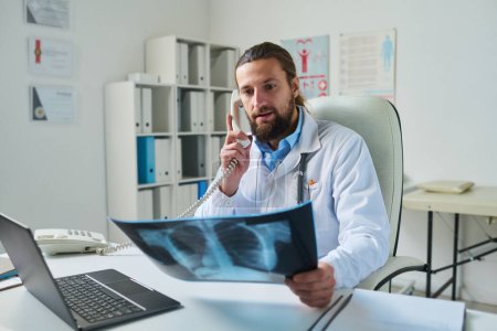Téléchargez les photos : Radiologiste confiant regardant la radiographie pulmonaire tout en étant assis par le lieu de travail dans le bureau médical et parlant au patient dans le récepteur téléphonique - en image libre de droit