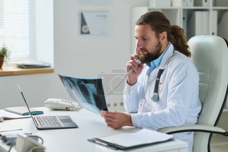 Téléchargez les photos : Clinicien masculin confiant en blouse de laboratoire regardant les résultats des rayons X du patient pendant la consultation téléphonique assis par le lieu de travail dans son bureau - en image libre de droit