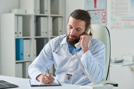 Foto de Joven médico con receptor de teléfono por oído haciendo notas en el documento médico mientras escucha al paciente durante la llamada telefónica - Imagen libre de derechos