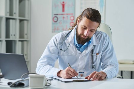 Téléchargez les photos : Jeune clinicien barbu en blouse de laboratoire prenant des notes dans un document médical assis sur le lieu de travail à l'hôpital - en image libre de droit