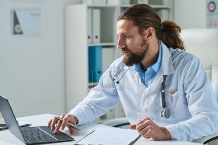 Téléchargez les photos : Jeune médecin généraliste masculin sérieux regardant écran d'ordinateur portable tout en étant assis par le lieu de travail dans les cliniques et en consultant des patients en ligne - en image libre de droit