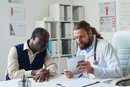 Photo for Young confident clinician with tablet making presentation of medical test results to patient while both sitting in clinical office and looking at screen - Royalty Free Image