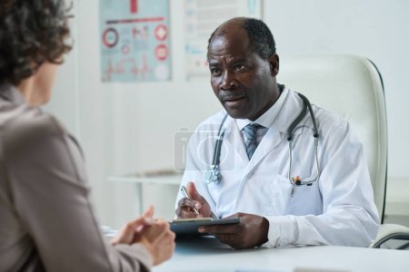 Téléchargez les photos : Clinicien afro-américain mature avec un document médical écoutant une patiente décrire ses symptômes pendant la consultation - en image libre de droit
