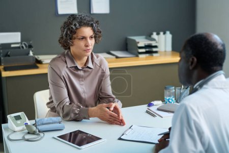 Téléchargez les photos : Patiente décrivant ses symptômes alors qu'elle était assise à son bureau devant un clinicien masculin afro-américain mature lors d'une consultation médicale - en image libre de droit