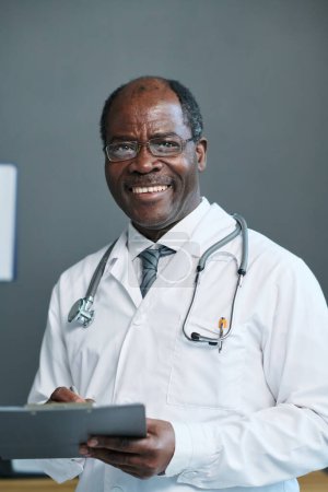 Téléchargez les photos : Médecin généraliste expérimenté avec succès avec un document médical regardant la caméra avec le sourire tout en faisant la prescription - en image libre de droit