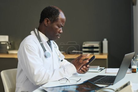 Téléchargez les photos : Vue latérale d'un clinicien mature sérieux défilant dans un smartphone assis devant un ordinateur portable par le lieu de travail et consultant des patients en ligne - en image libre de droit