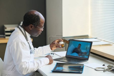 Foto de Médico general afroamericano maduro recomendando píldoras al paciente masculino enfermo en línea en la pantalla del ordenador portátil durante la consulta - Imagen libre de derechos