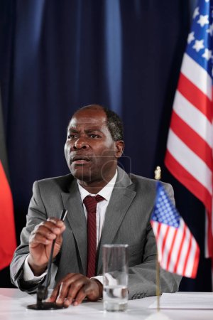 Foto de Hombre afroamericano maduro orador o político en ropa formal hablando en micrófono mientras participa en una conferencia o cumbre - Imagen libre de derechos