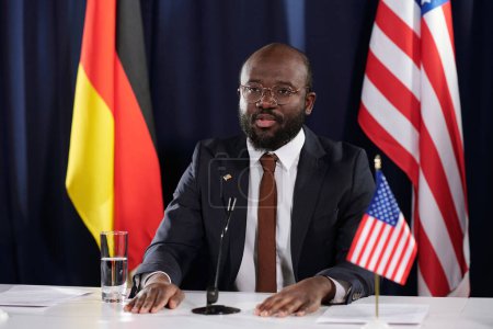 Foto de Joven representante varón afroamericano confiado de EE.UU. que usa traje haciendo discurso mientras está sentado frente al micrófono en la cumbre - Imagen libre de derechos