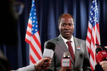 Photo for Mature male deputy in suit looking at one of journalists during interview at press conference while standing between two American flags - Royalty Free Image