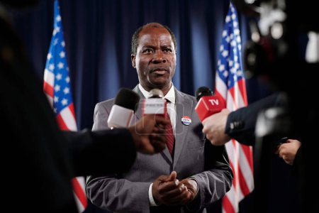 Téléchargez les photos : Homme politique afro-américain mature et confiant, en tenue de cérémonie, posant des questions aux journalistes tout en parlant dans des microphones - en image libre de droit