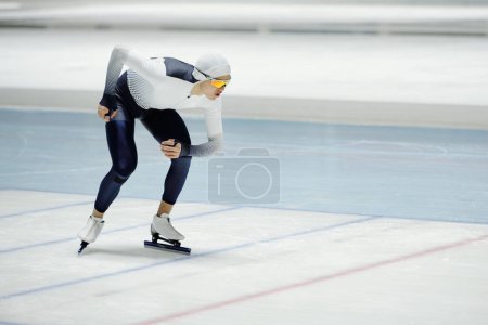 Téléchargez les photos : Jeune sportif en uniforme de patinage de vitesse et lunettes glissant sur patinoire à l'intérieur de la zone moderne tout en prenant part à la compétition - en image libre de droit