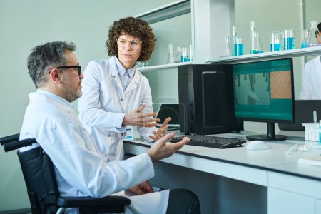 Confident mature female virologist talking to male colleague with disability sitting in wheelchair in front of computer during discussion