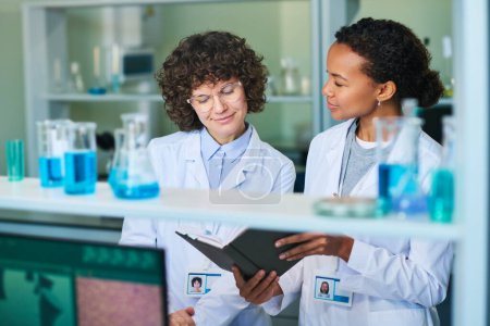 Téléchargez les photos : Jeune chercheuse brune en blouse de laboratoire et lunettes regardant les notes dans un carnet tenu par un collègue afro-américain - en image libre de droit