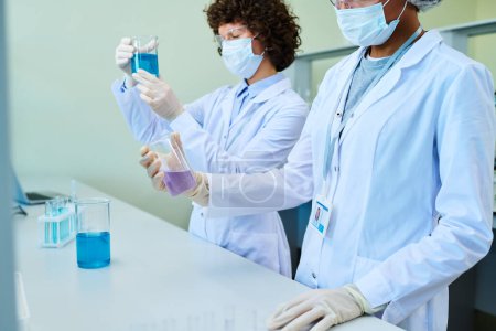 Photo for Two young intercultural female clinicians in lab coats and protective masks working with liquids in test tubes while standing by workplace - Royalty Free Image