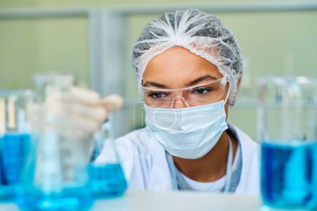 Foto de Enfoque en la cara de una joven química afroamericana que toma un tubo de ensayo con líquido azul mientras realiza un experimento científico en laboratorio - Imagen libre de derechos